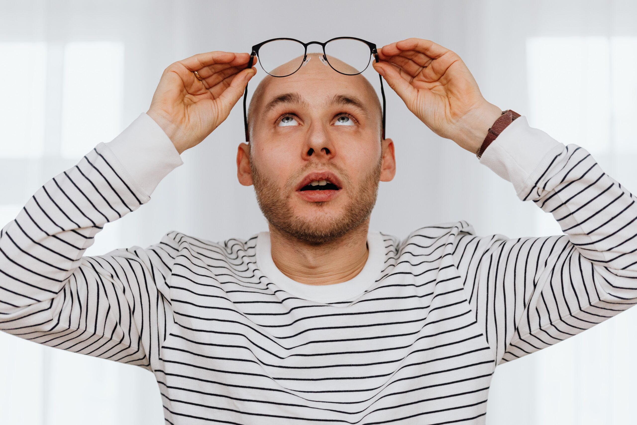 A bald man is looking carefully at his eyeglasses as he holds them above his head