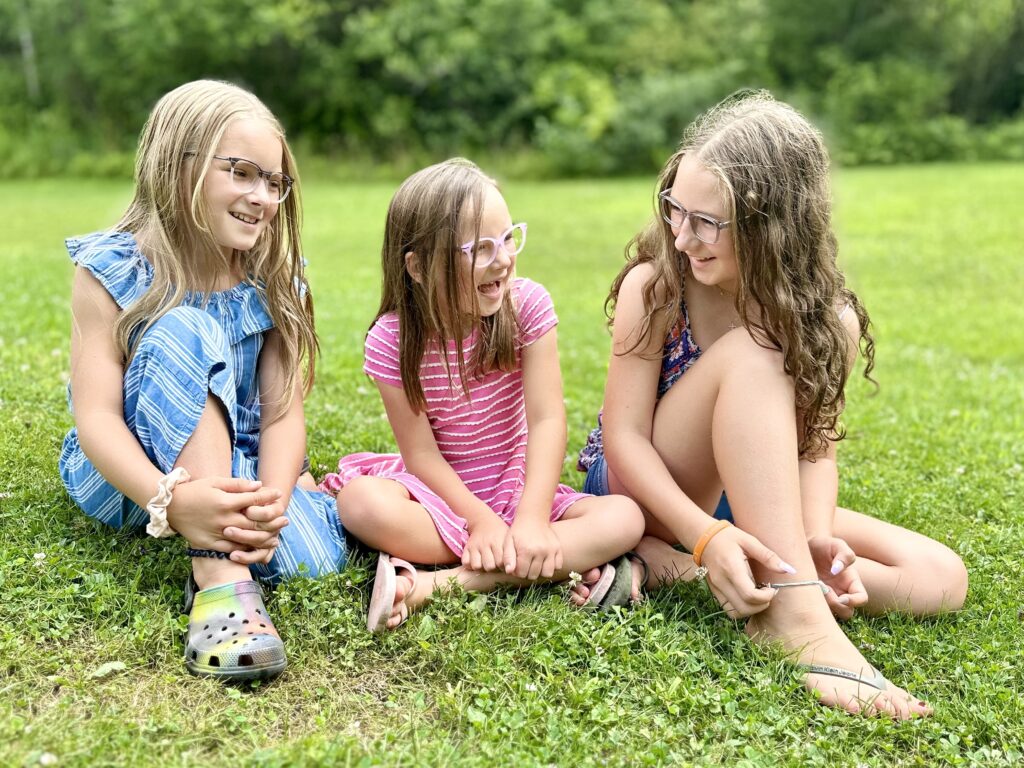 Three youn girls sit together on the grass. Each are wearing eyeglasses and smiling. 